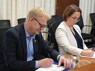 Nahaufnahme: Ein Mann und eine Frau sitzen am Tisch in einem Sitzungssaal. beide notieren sich etwas auf Papier. Sie wirken offiziell, der Mann trägt Anzug, sie einen weißen Blazer.