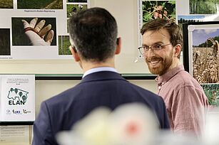 Zwei Männer im Gespräch vor einer Posterwand mit landwirtschaftlichen Motiven. Der eine Mann wendet uns den Rücken zu, der ander erklärt diesem lächelnd etwas. Wir können ihn von der Seite sehen.